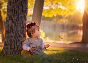 Child sits by tree in autumn park at sunset as golden sunlight filters through leaves and butterfly floats nearby.