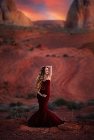 Figure in a flowing red velvet gown poses dramatically against red rock formations at sunset in the desert.