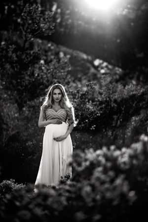 A moody black and white maternity portrait shows an expectant mother in a flowing white dress standing in garden scenery.