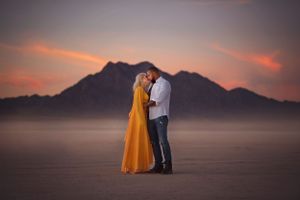 A couple in elegant attire embraces on a desert plain against a dramatic mountain silhouette at sunset.