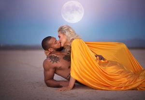 A romantic couple poses on sand dunes at dusk with a dramatic full moon in a purple-blue sky as backdrop.