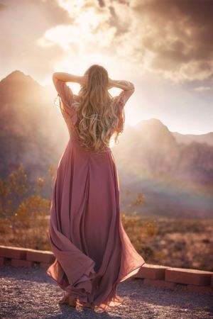 A long-haired figure in a flowing mauve dress stands against a mountainous landscape at sunset.
