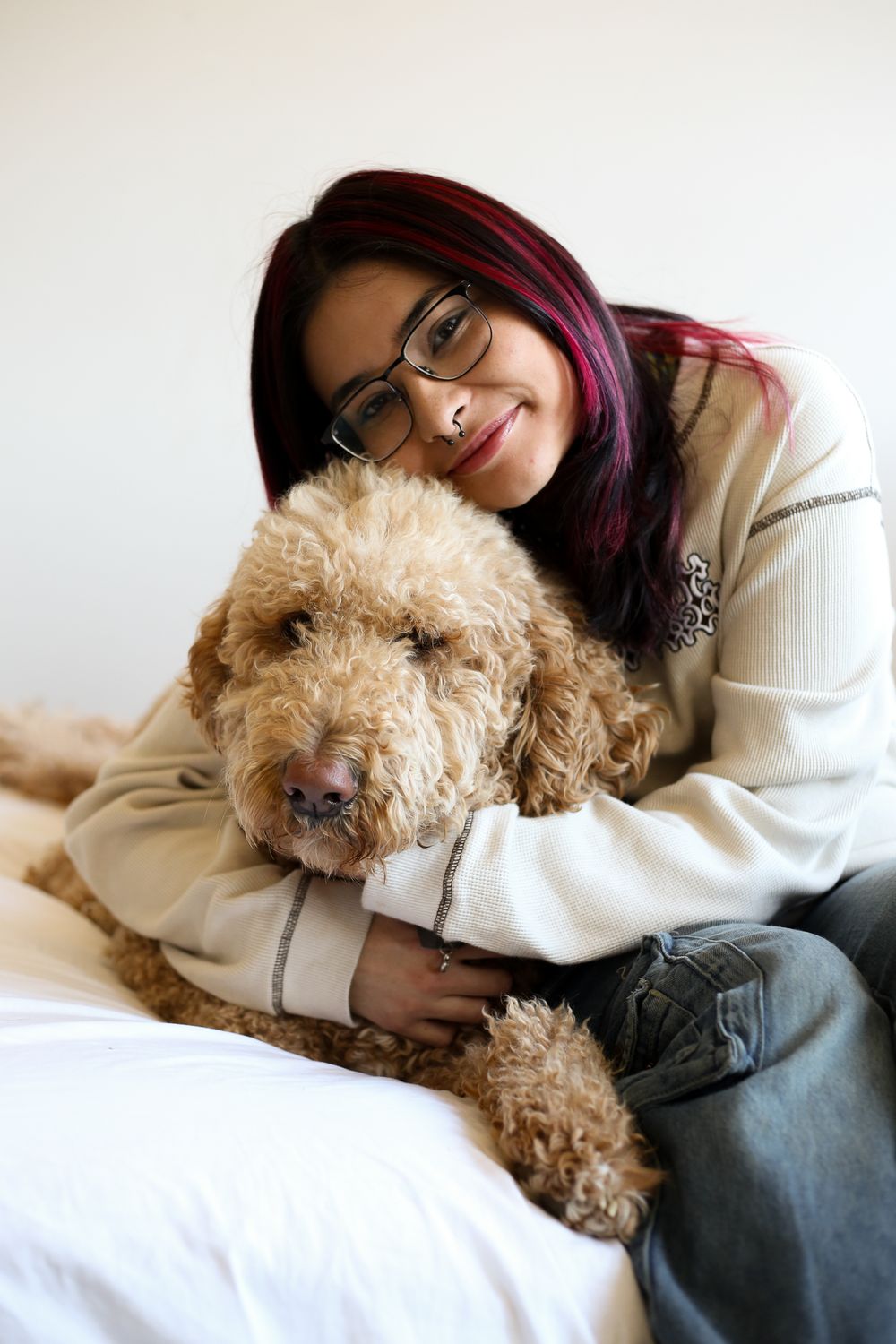 A person with purple hair cuddles with a fluffy goldendoodle dog on a white bed.