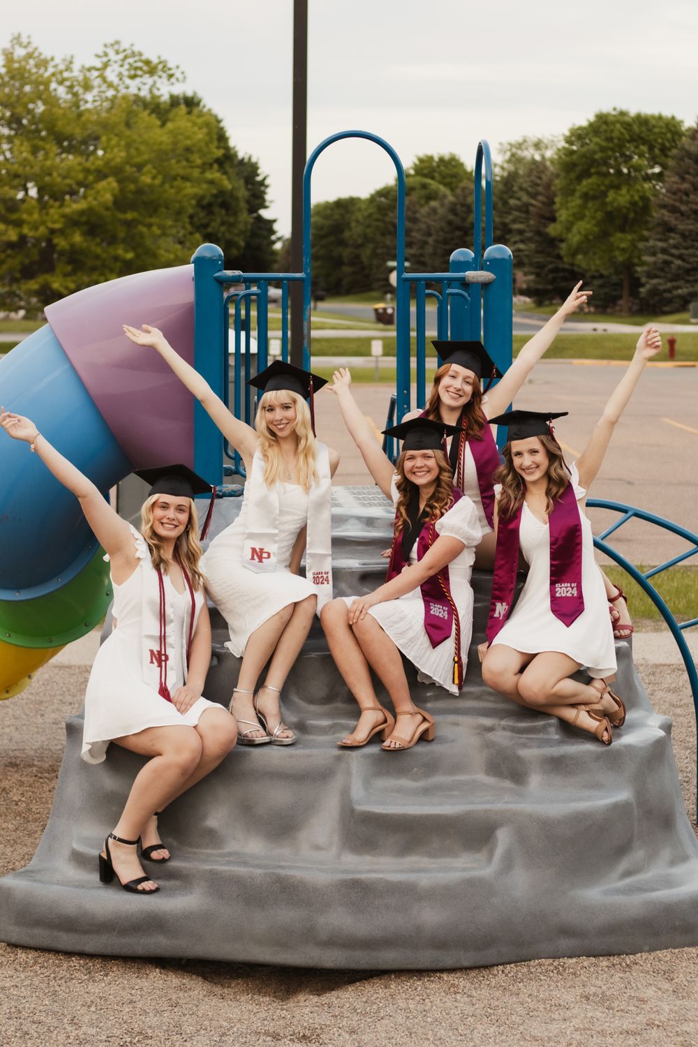 Group of graduates in white dresses and burgundy stoles posing on playground equipment.