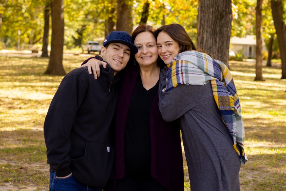 Three people embracing and smiling together in a park with golden autumn foliage in the background.