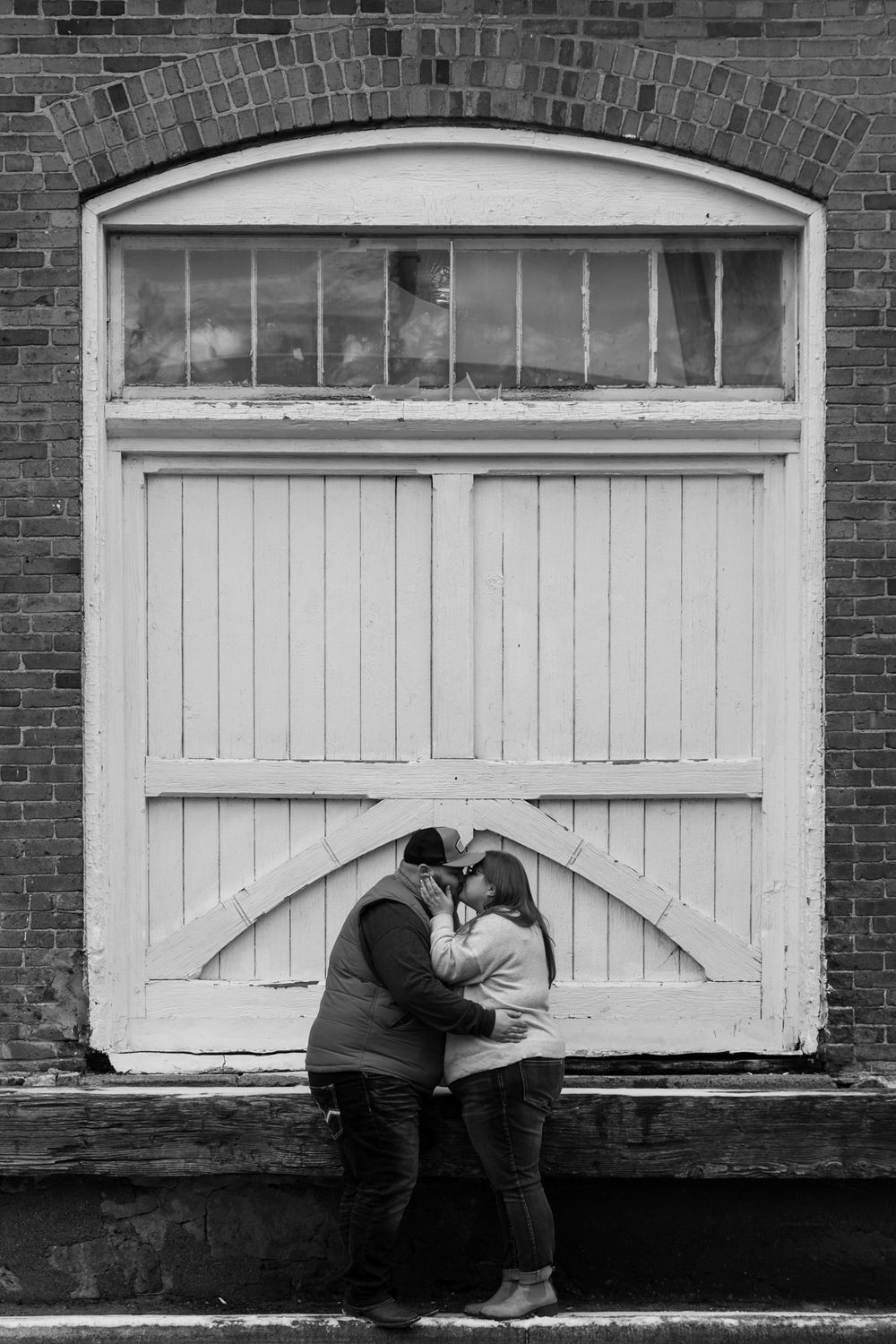 Couple sharing an intimate moment against a white wooden door with brick wall background in black and white.