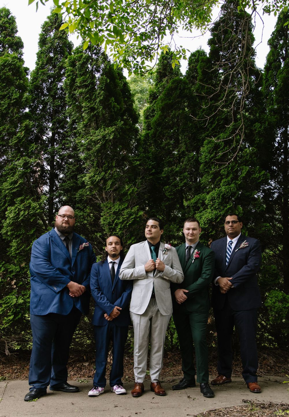 A group in formal attire poses in a creative line formation against a backdrop of lush green trees and foliage.