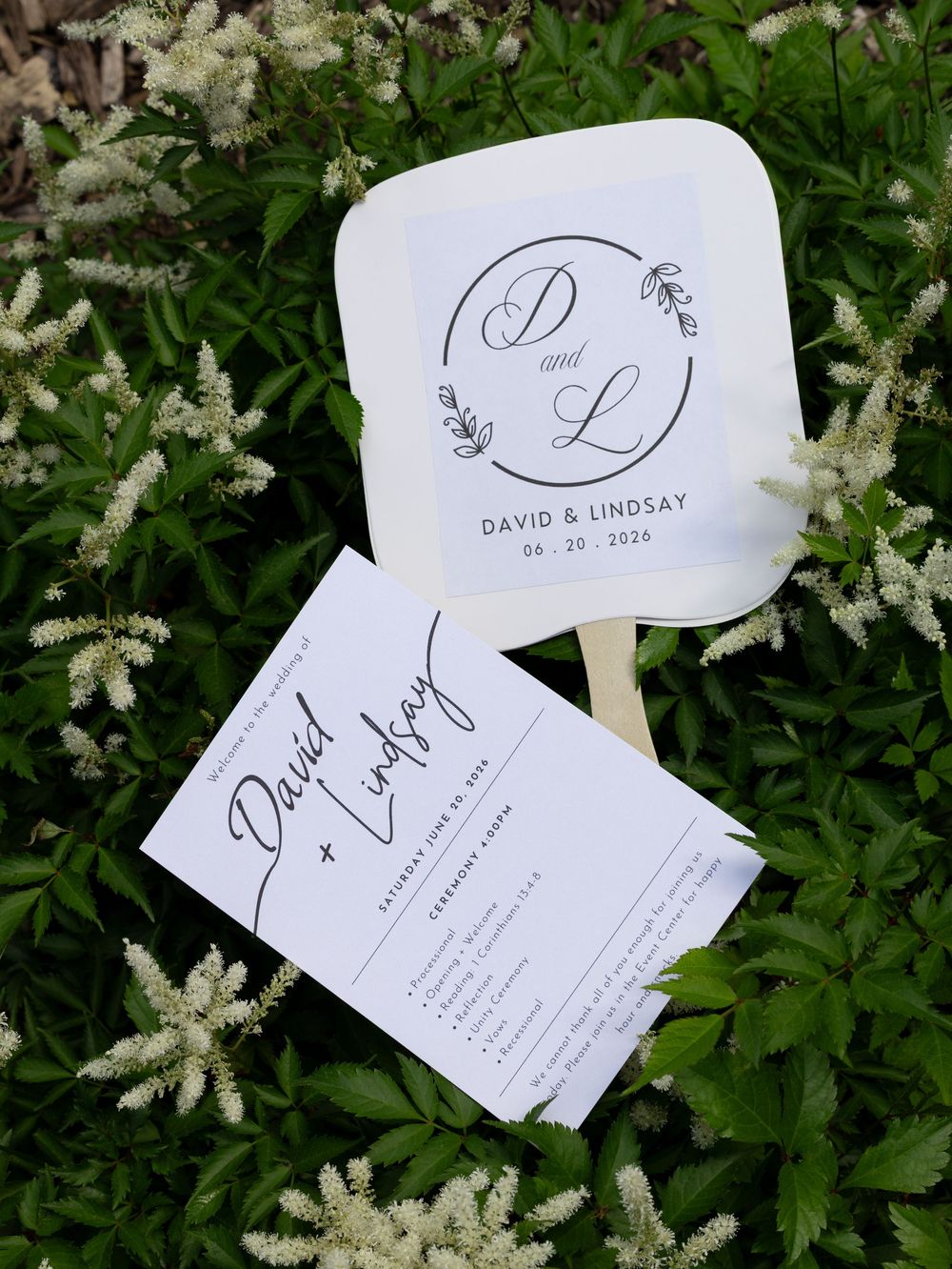 Wedding fan programs for David & Lindsay dated 08.20.2026, displayed on lush green foliage with white flowers.