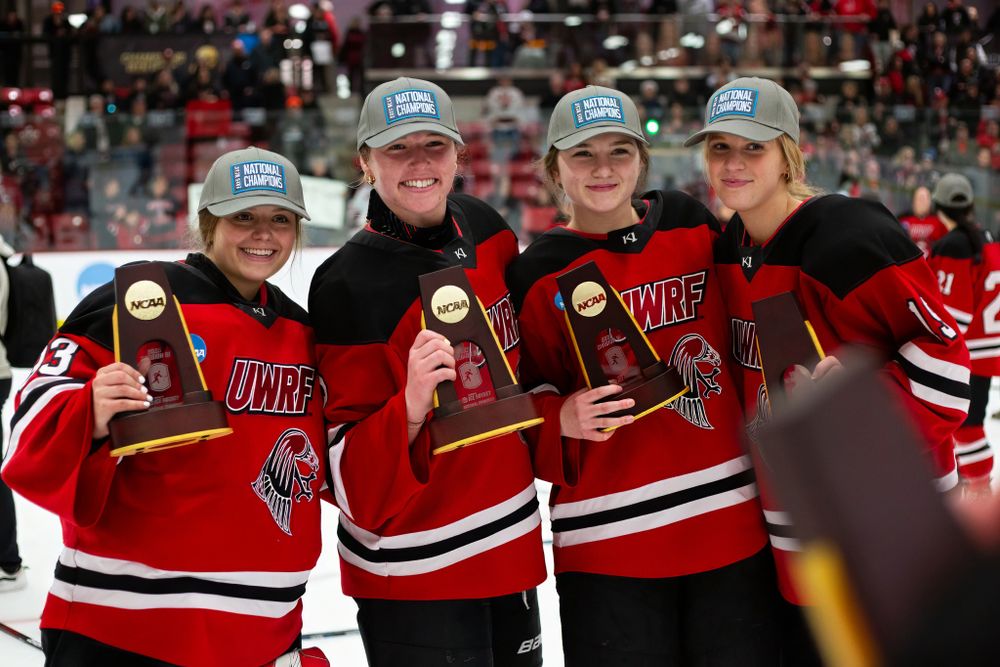 Four hockey players in red jerseys celebrate with championship trophies on the ice.