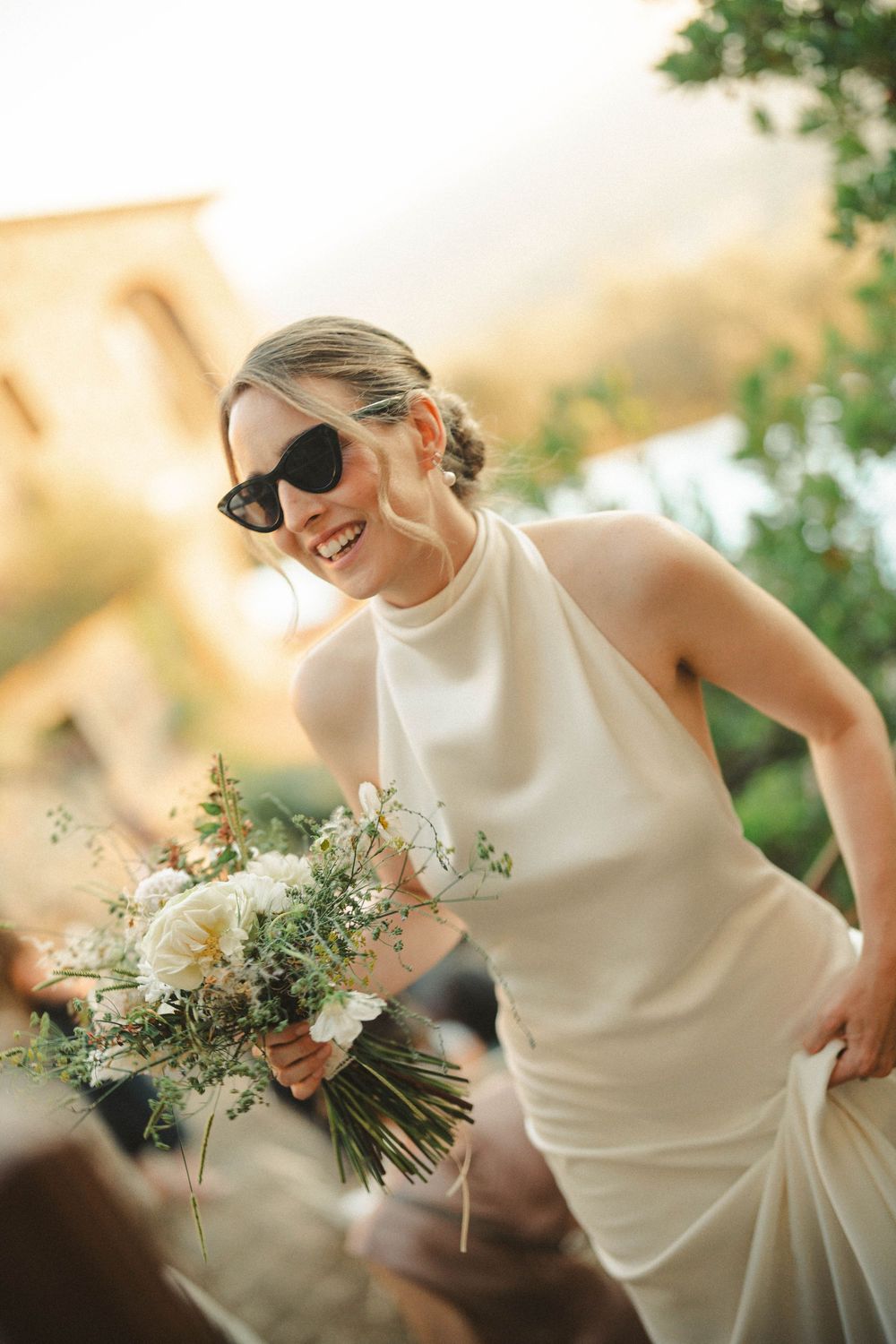 Joyful bride in sunglasses holds white and green bouquet in natural sunlight.