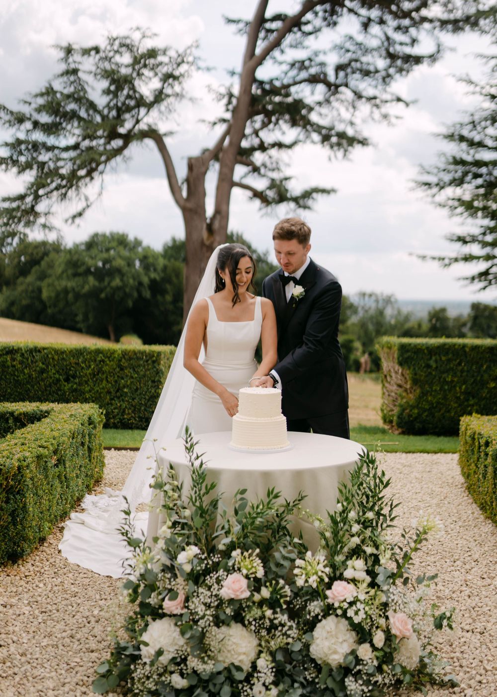 Wedding couple cutting cake in a beautiful garden setting with white floral arrangements.