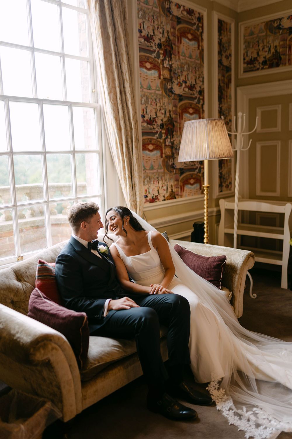 Romantic wedding scene in elegant room with floral wallpaper and large window.