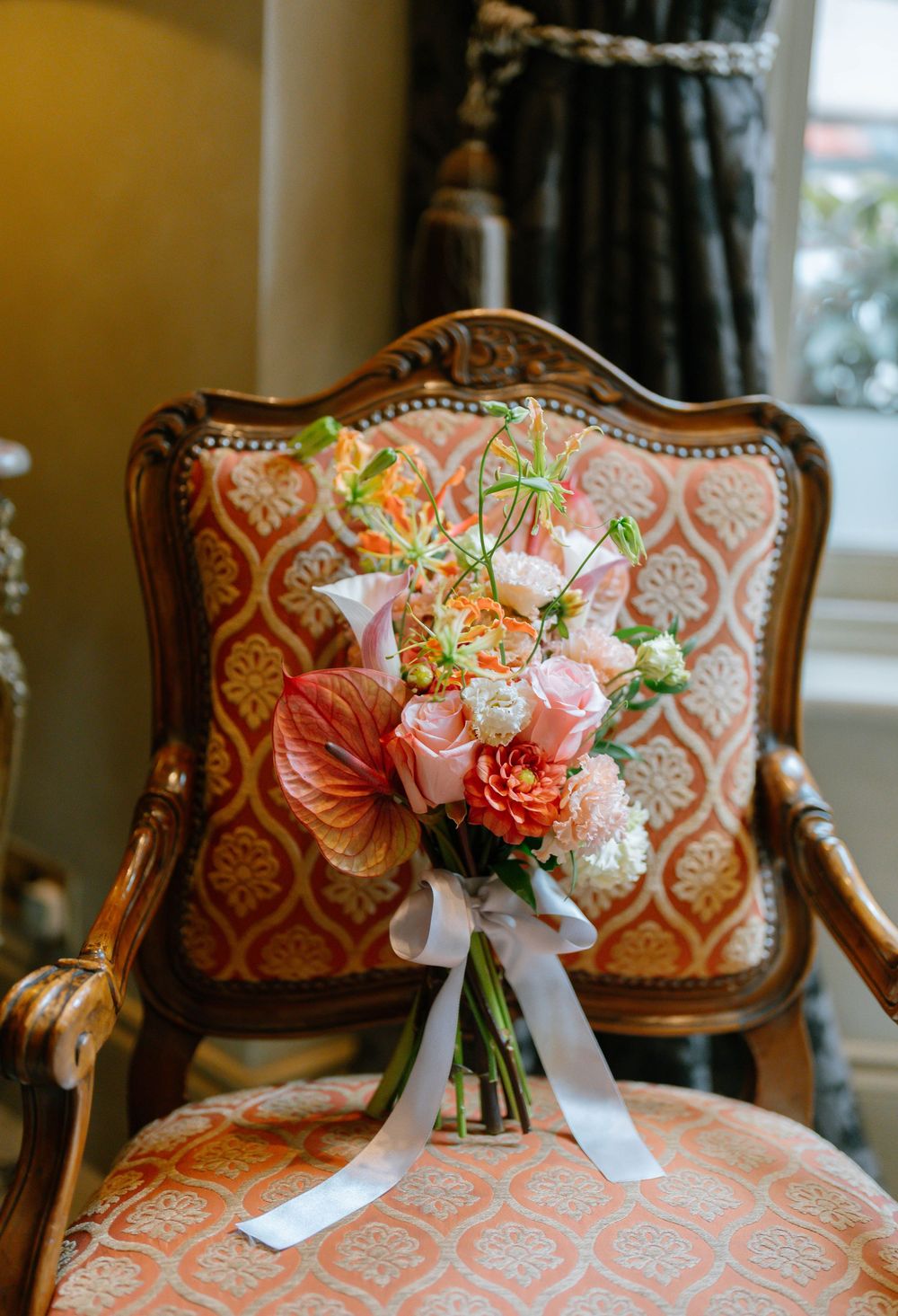 Elegant bridal bouquet with pink and coral flowers resting on an antique chair with patterned upholstery.