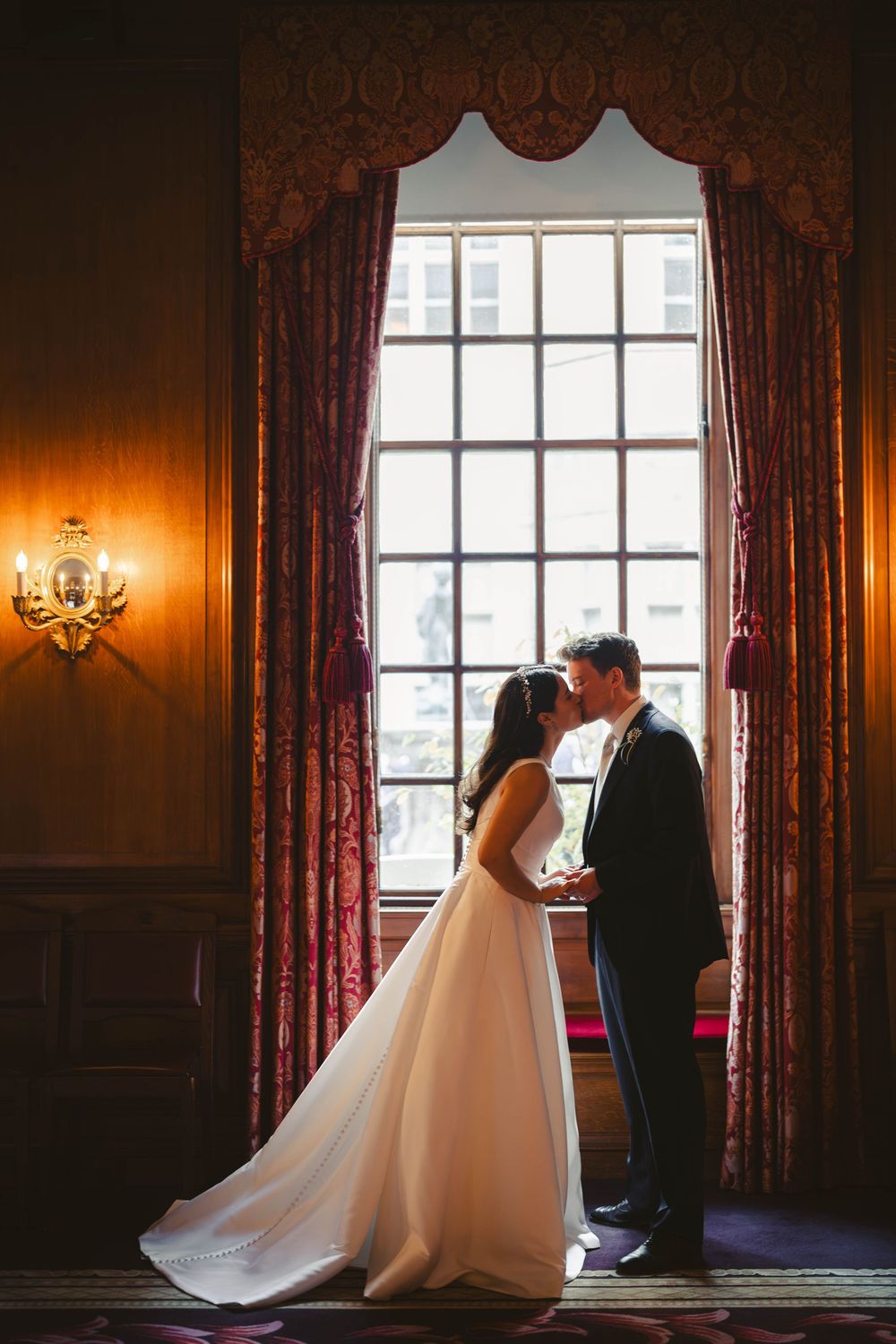 A tender moment between newlyweds silhouetted against a grand window in a warmly lit historic interior.