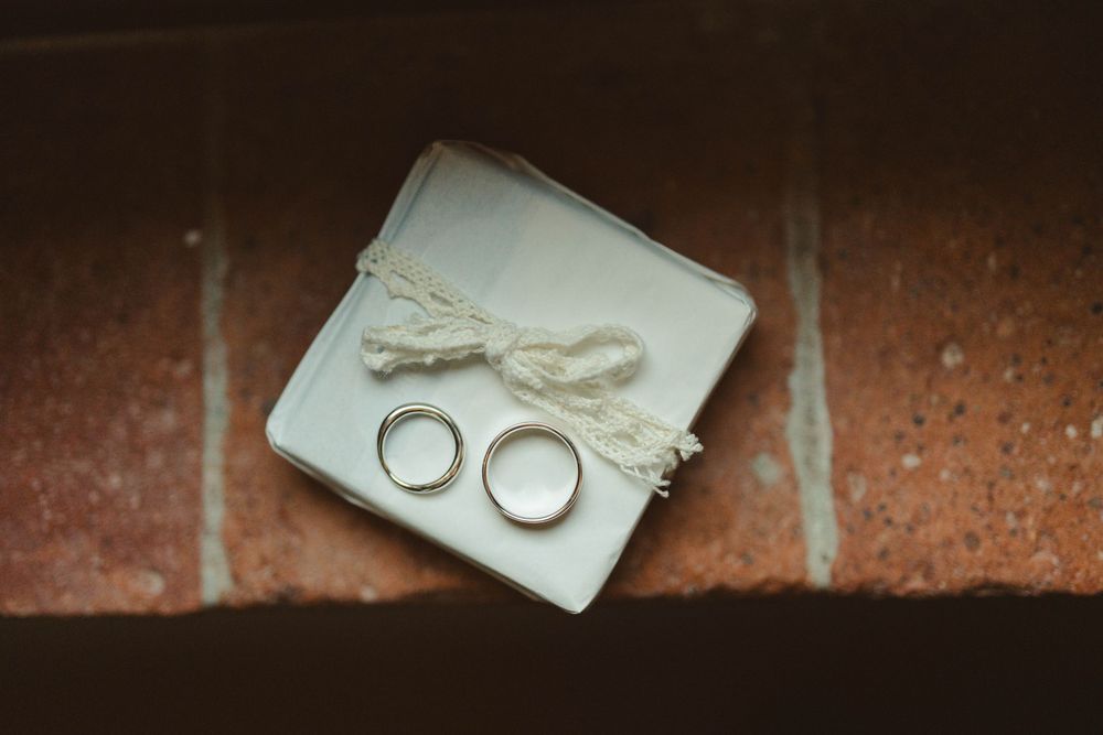 Two wedding rings rest on a white cushion with delicate string detail against a rustic brick background.
