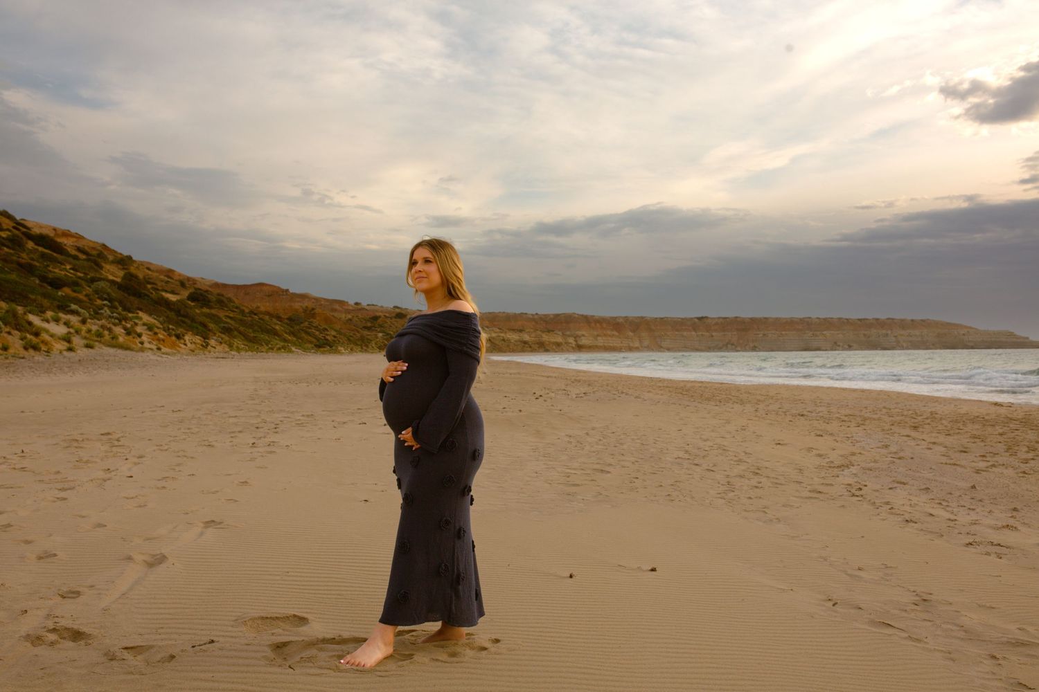 Woman in dark dress standing alone on beach during cloudy sunset.