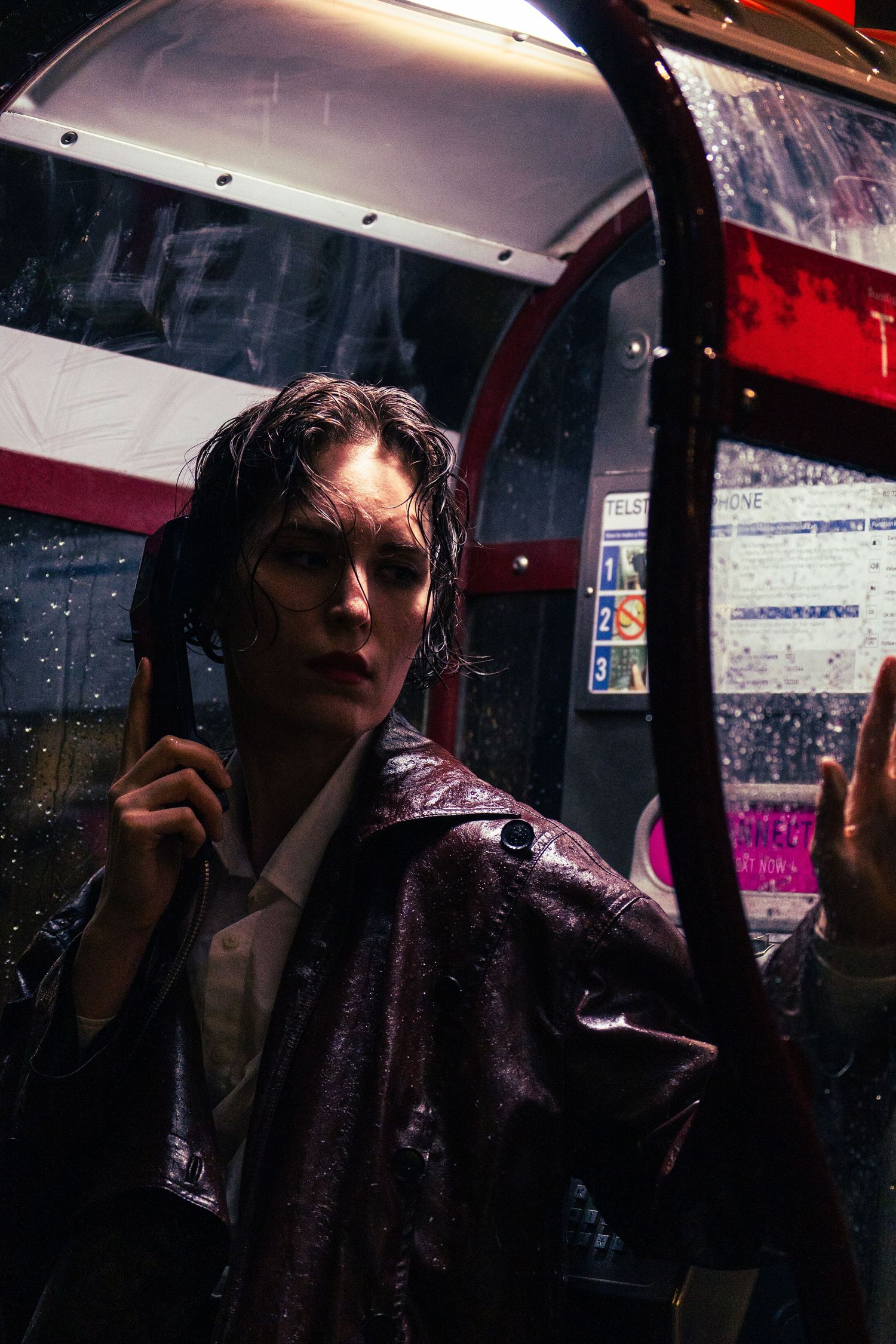 Young person in shiny purple jacket stands in rain at a bus stop with wet hair and dramatic lighting.