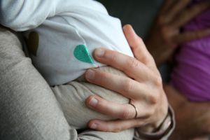 Close-up of hand with wedding ring resting on white clothing with small teal heart detail.