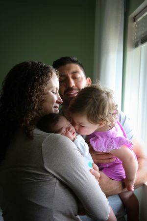 Intimate family moment by window as they embrace and cuddle together in natural light.