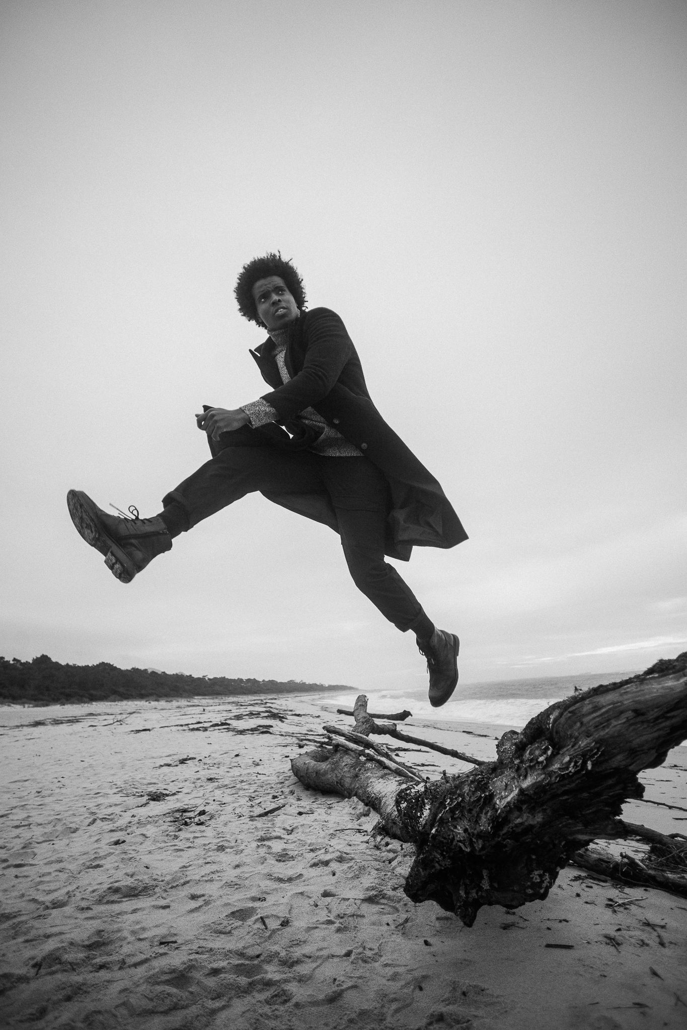 Dynamic black and white photo of someone jumping over driftwood on a beach.