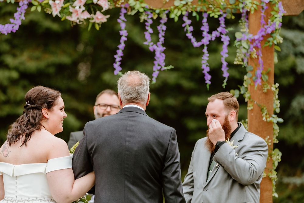 Outdoor wedding ceremony with bride, groom wiping tears, and officiant under a floral arch with purple wisteria.