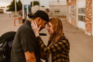 A tender moment between two people in a plaid shirt and black hat standing close on a quiet street at sunset.