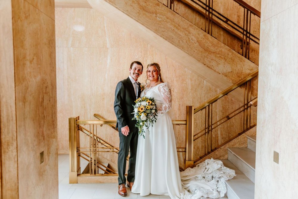 Bride and groom smiling together on marble staircase, bride holding floral bouquet in elegant white gown.
