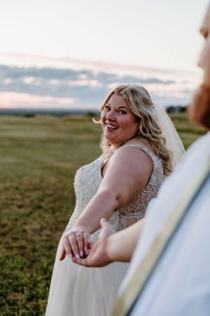 Smiling bride in lace wedding dress and veil holds groom's hands in golden hour field.