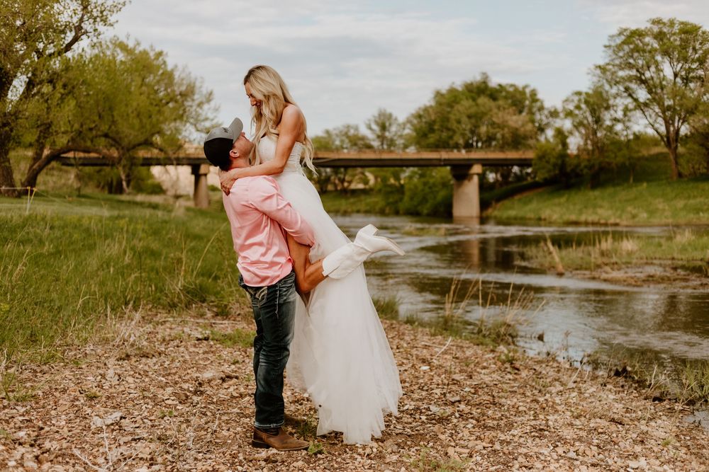 A bride in a white dress and young boy share tender moments along a riverbank in a rustic outdoor setting.