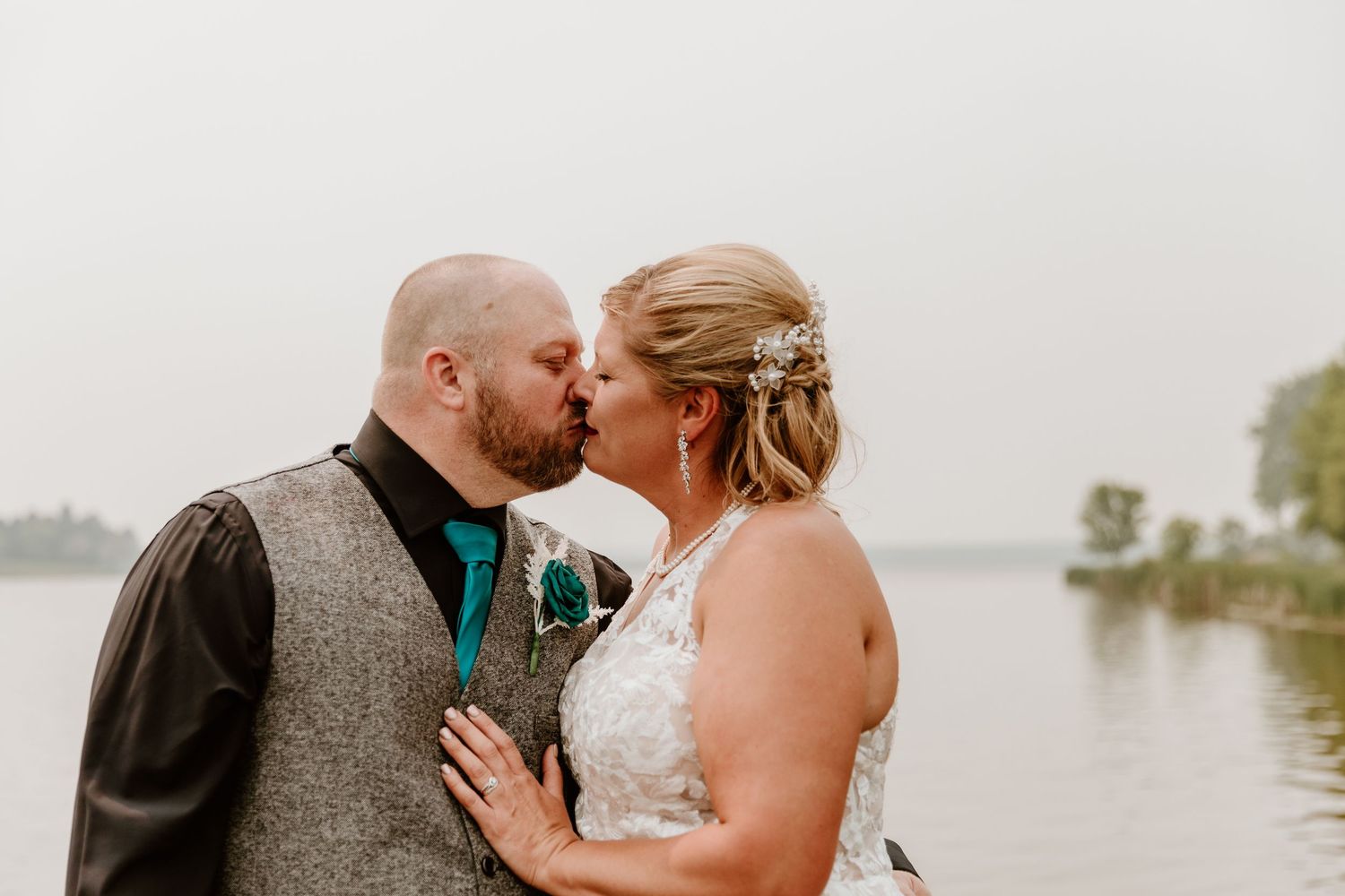 Bride and groom sharing a romantic kiss near a serene lake, with the bride in a lace gown and groom in a grey vest.