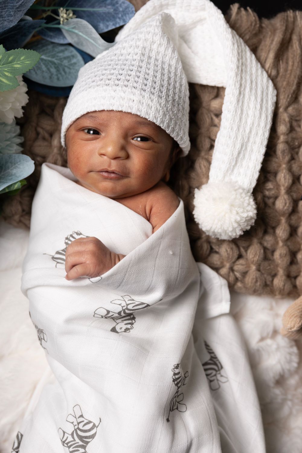 An infant wearing a white knit hat with pom pom and swaddled in a printed white blanket.