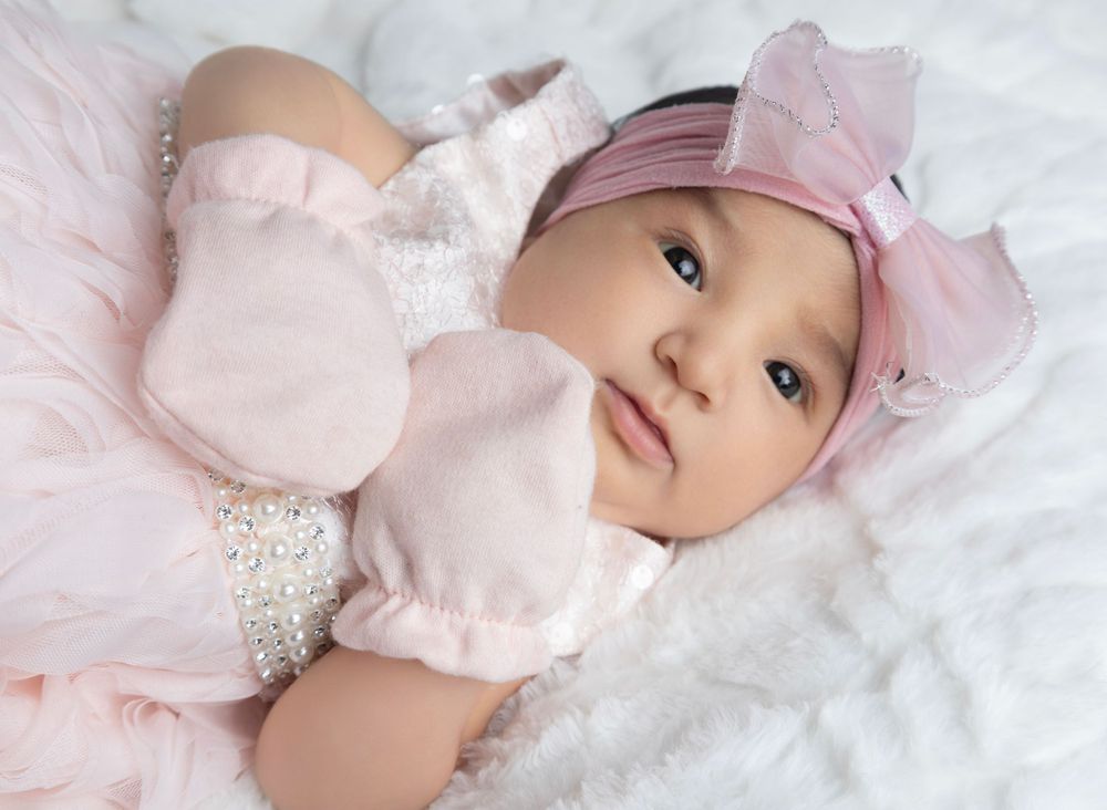 Baby in soft pink outfit with matching headband rests on white lacy background.