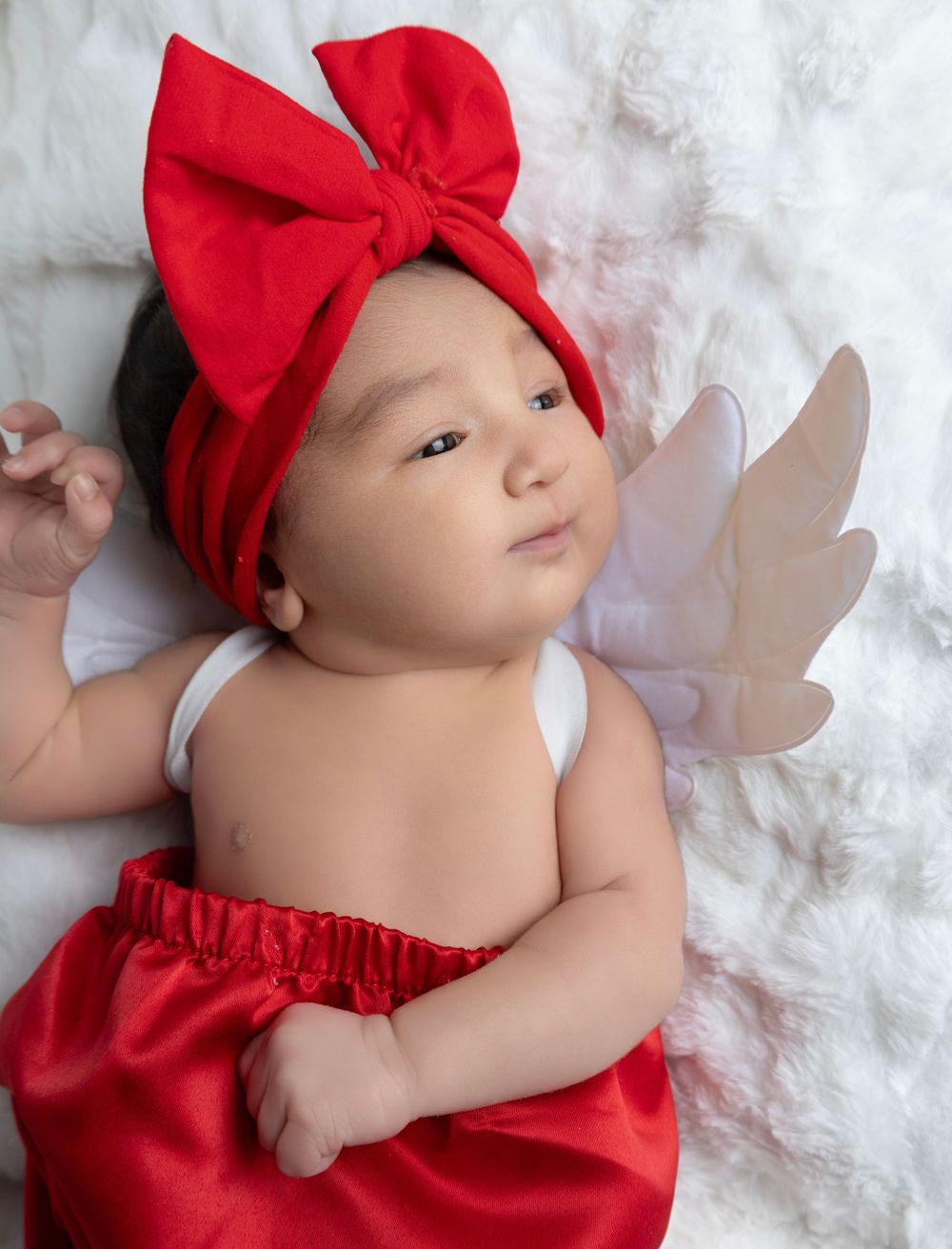 Baby wearing red bow headband and skirt lying on white furry backdrop with small decorative wings.