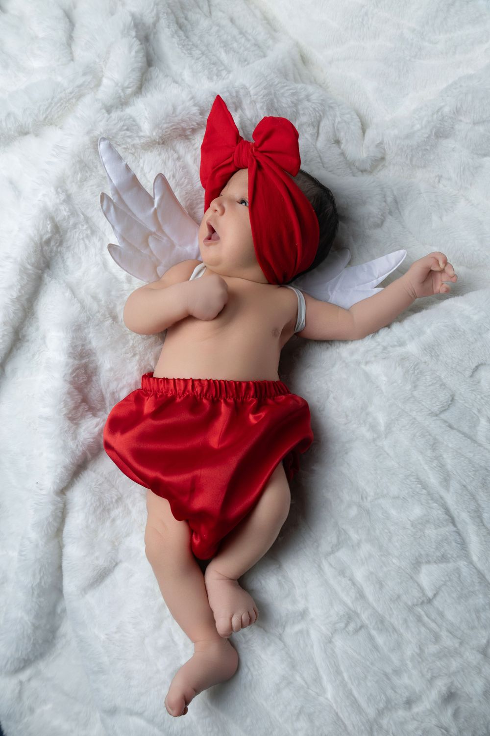 Baby wearing red headband and bloomers lies peacefully on soft white blanket with arms spread out.