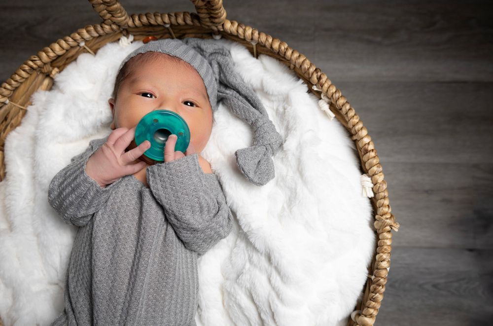 Baby in grey knit clothing with teal pacifier resting in round woven basket.