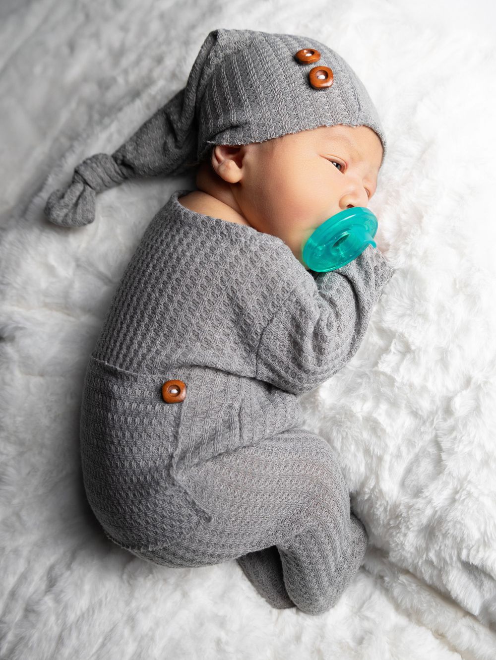 Sleeping baby wearing grey knit outfit with teal pacifier and decorative buttons.