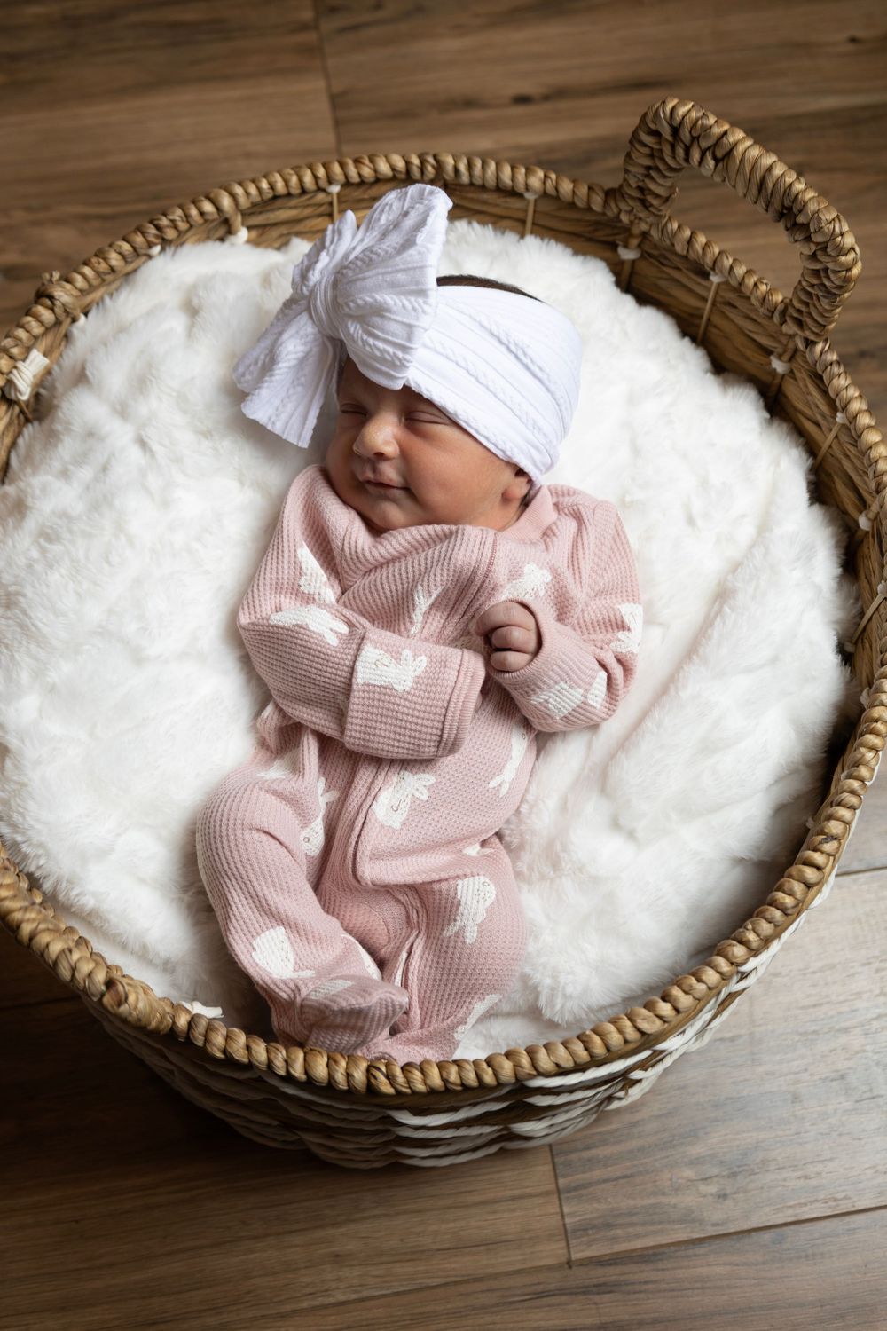 Newborn baby wearing soft pink outfit and white bow headband sleeping peacefully in wicker basket with white blanket.