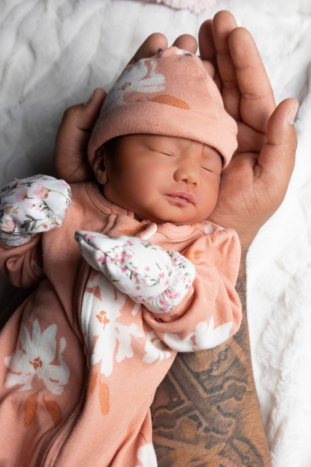 A sleeping newborn in a pink outfit and hat rests gently in an adult's tattooed hand on white bedding.