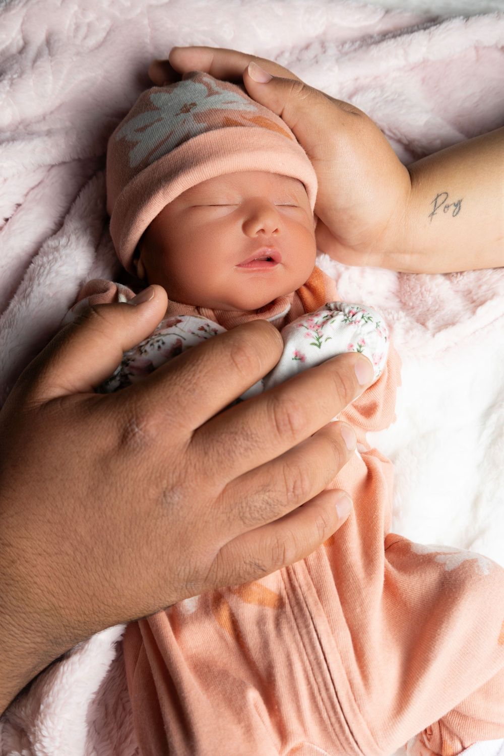 A sleeping newborn wearing a soft pink hat rests peacefully while being cradled by gentle hands on white blankets.