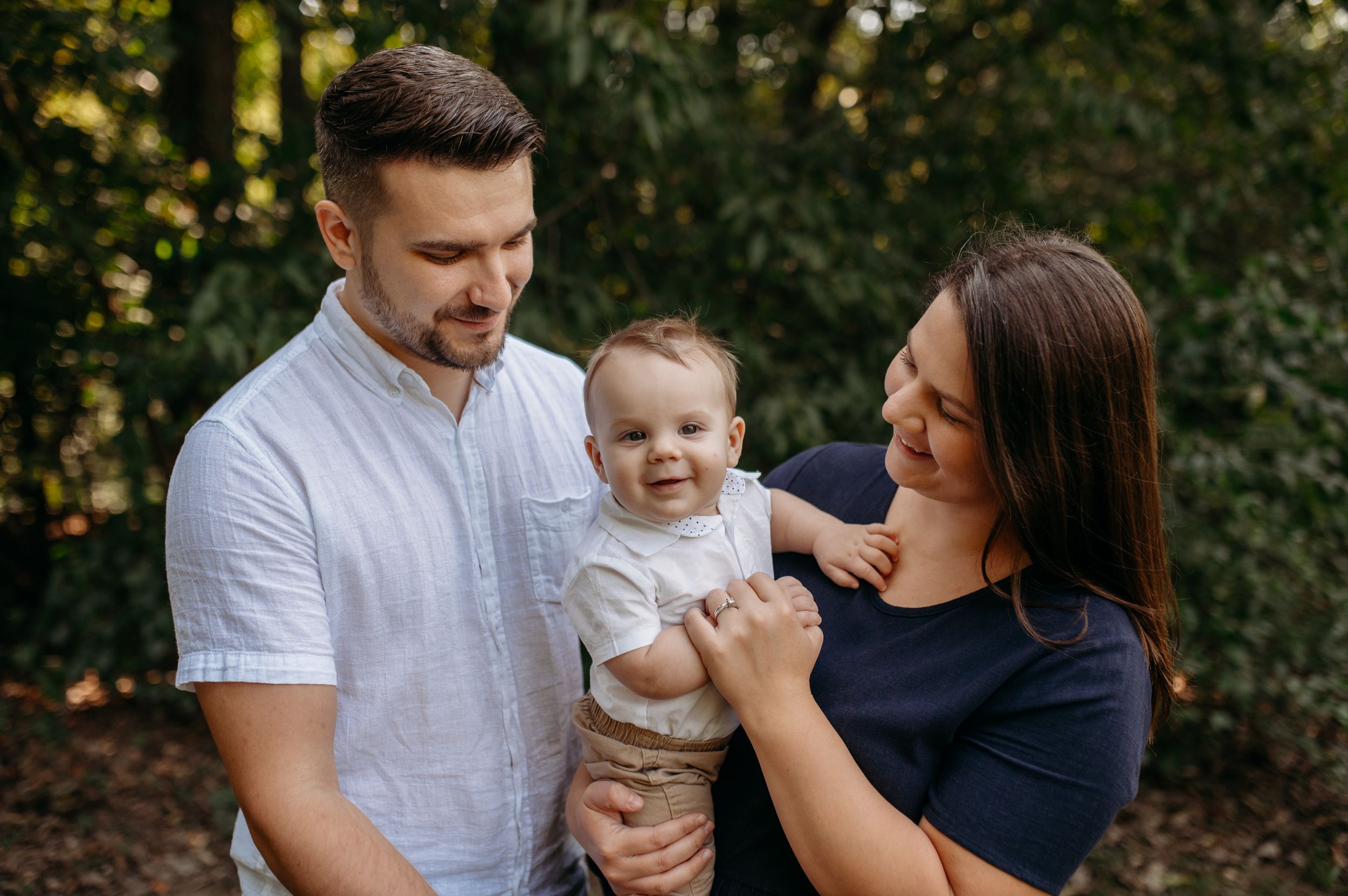 Young parents look at their toddler, snuggled in their arms, as they all pose for family photos in a warm park in St. Charles county, MO.