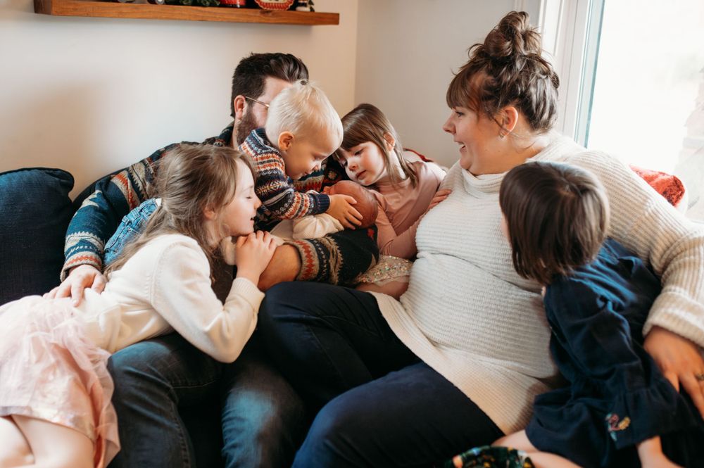 Mom and dad and their five children snuggle up on the couch all smiles and giggles as they pile on each other to kiss their new baby brother