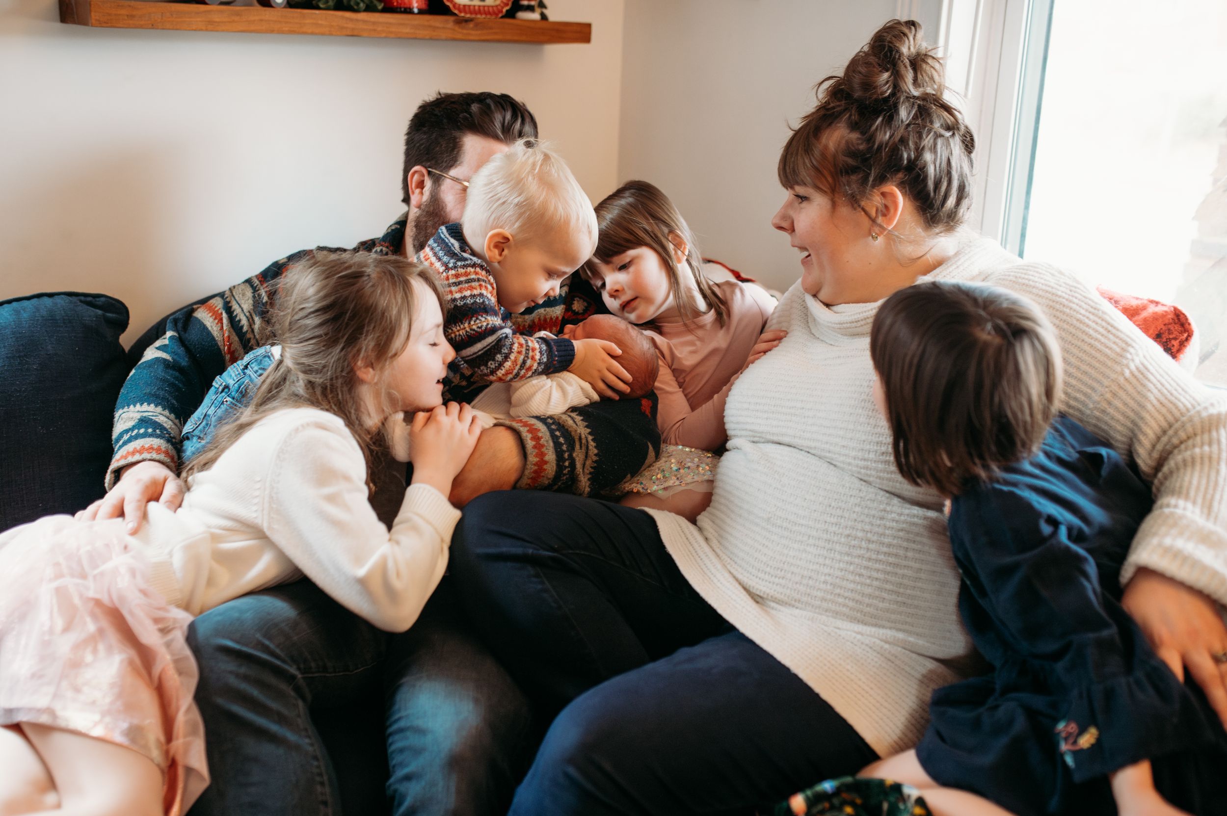 Mom and dad and their five children snuggle up on the couch all smiles and giggles as they pile on each other to kiss their new baby brother