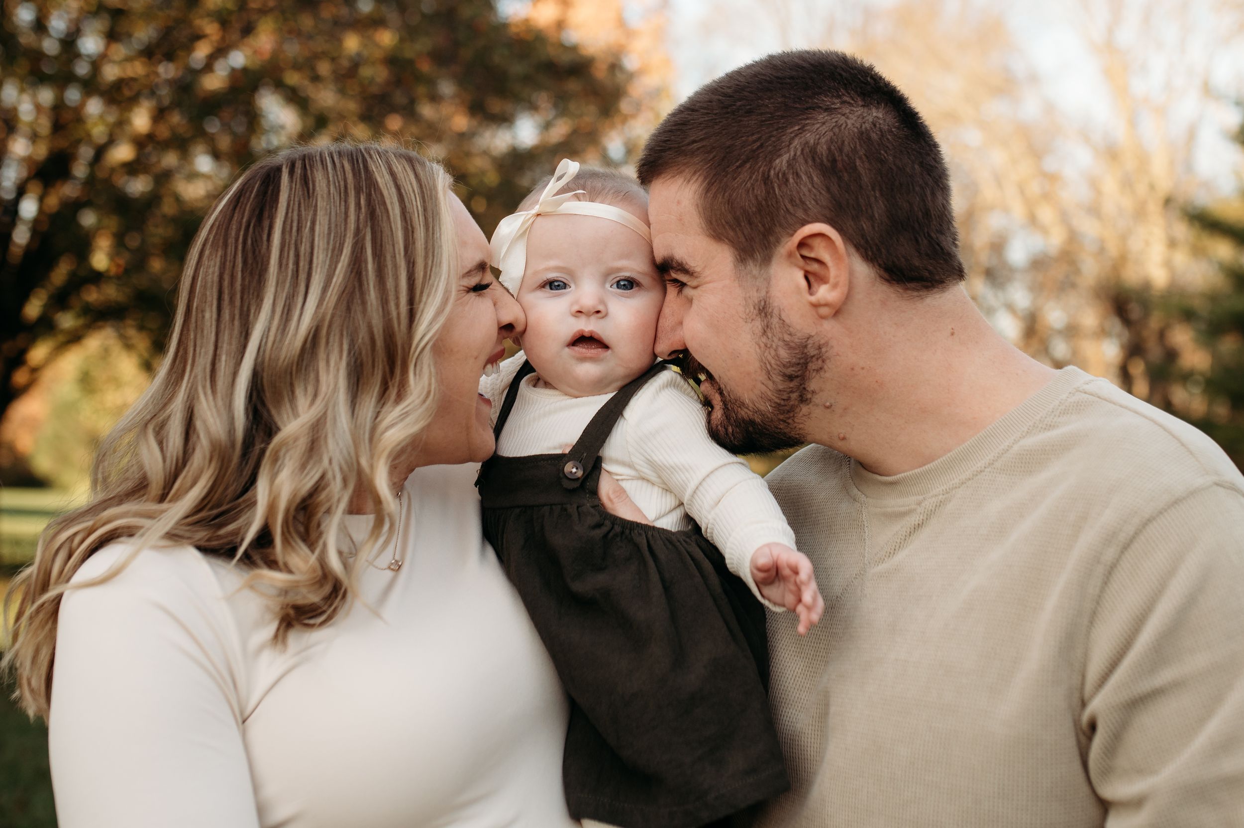 Mom and Dad kiss both sides of baby girl's face as she looks directly ahead and smiles at the camera for their first family photos