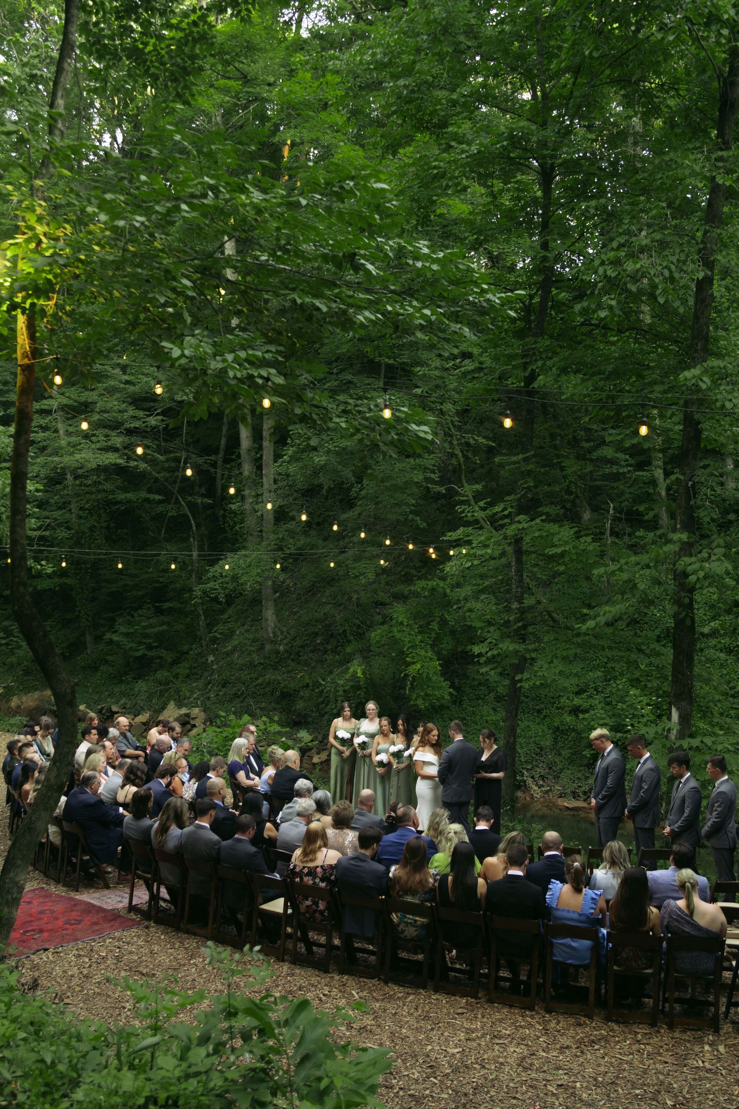 Outdoor forest wedding ceremony with string lights and guests seated on wooden chairs. Kentucky. Cave wedding. Documentary photographer
