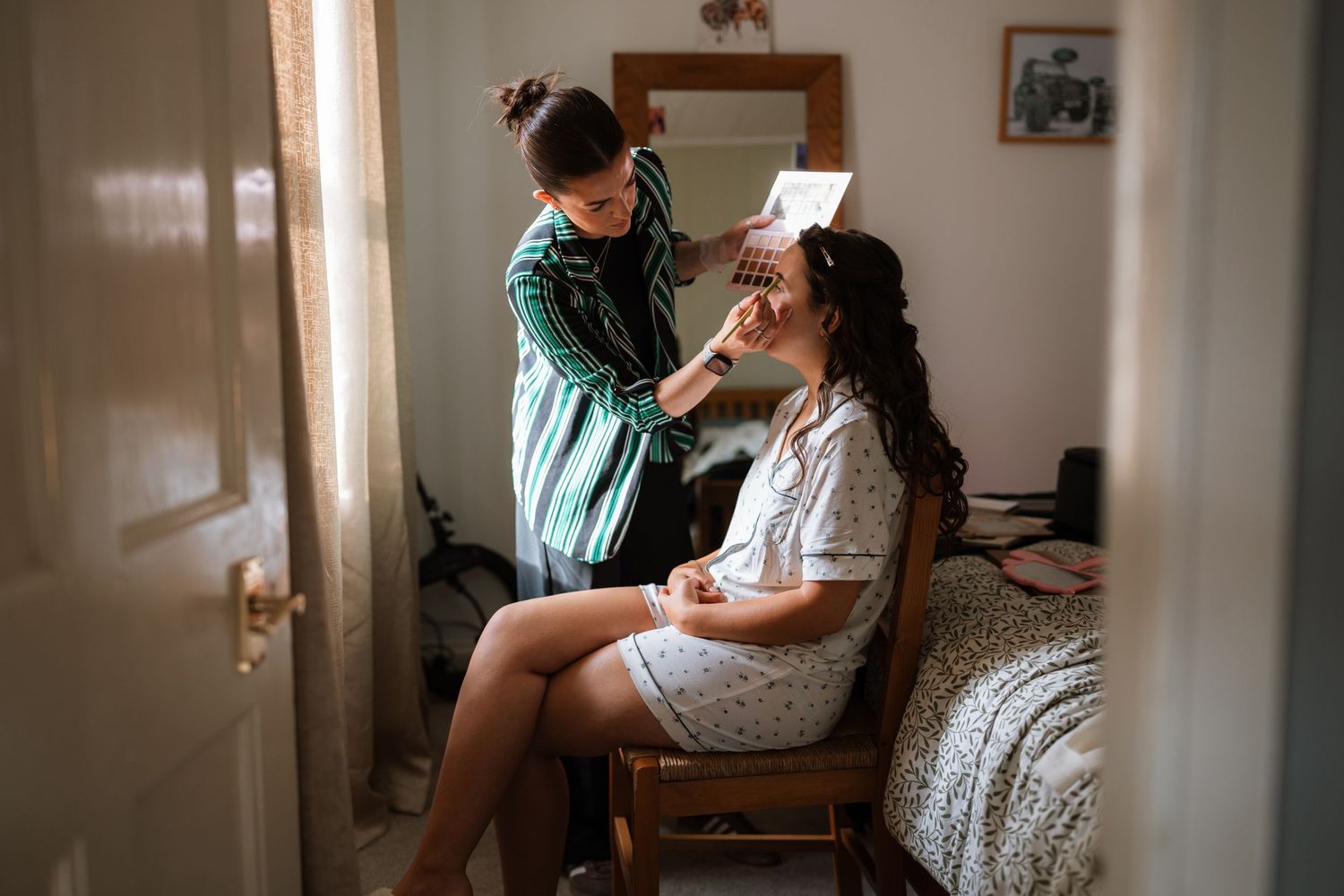 Makeup artist in striped shirt applies cosmetics to seated client in navy blue top during beauty session.