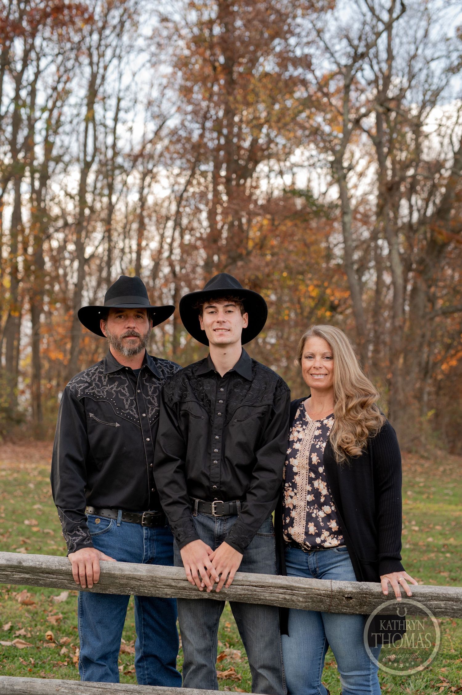 Family leaning on wooden fence in western attire with fall foliage in background.