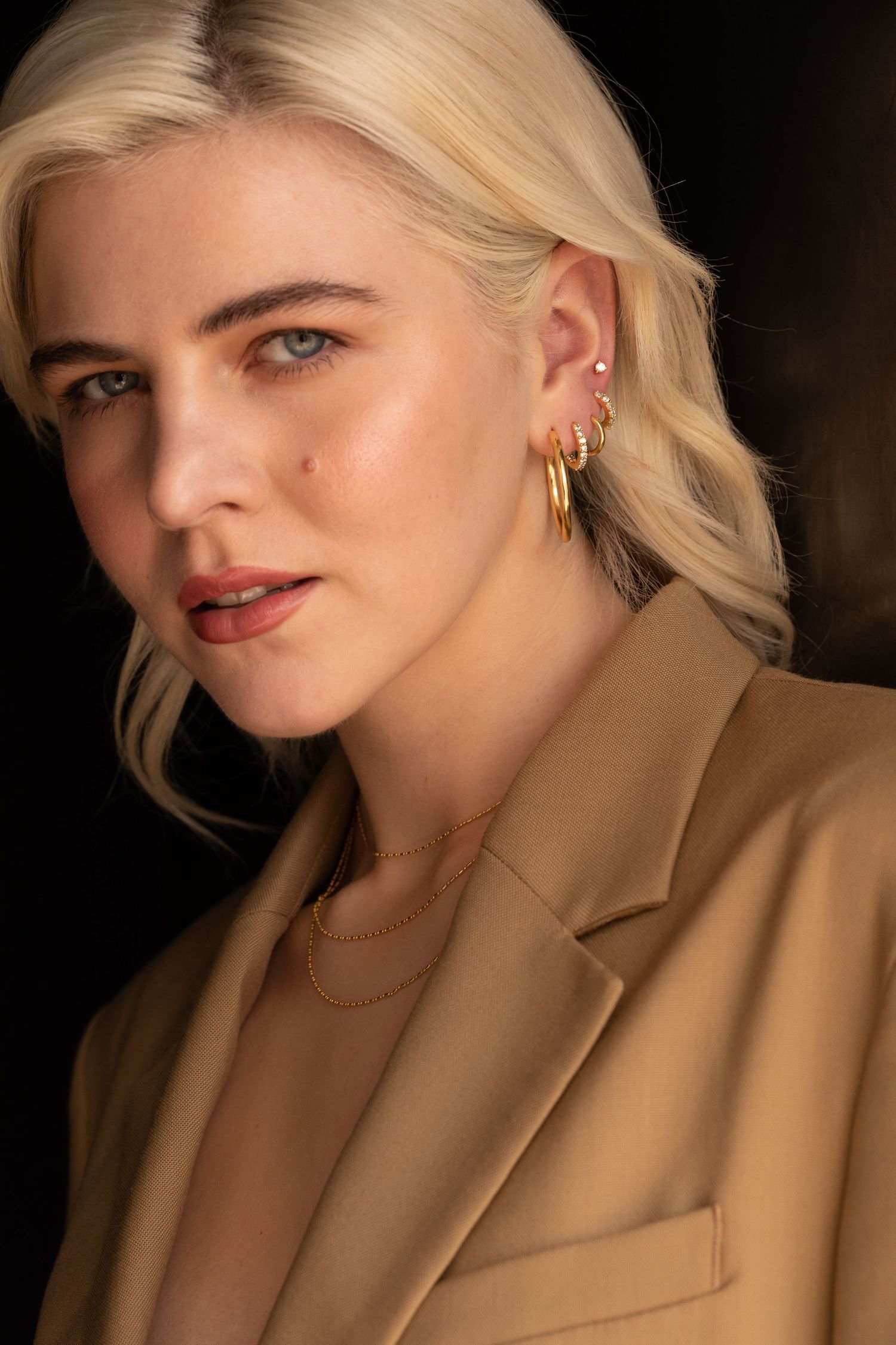 Portrait shot featuring gold jewelry and camel blazer in dramatic lighting.