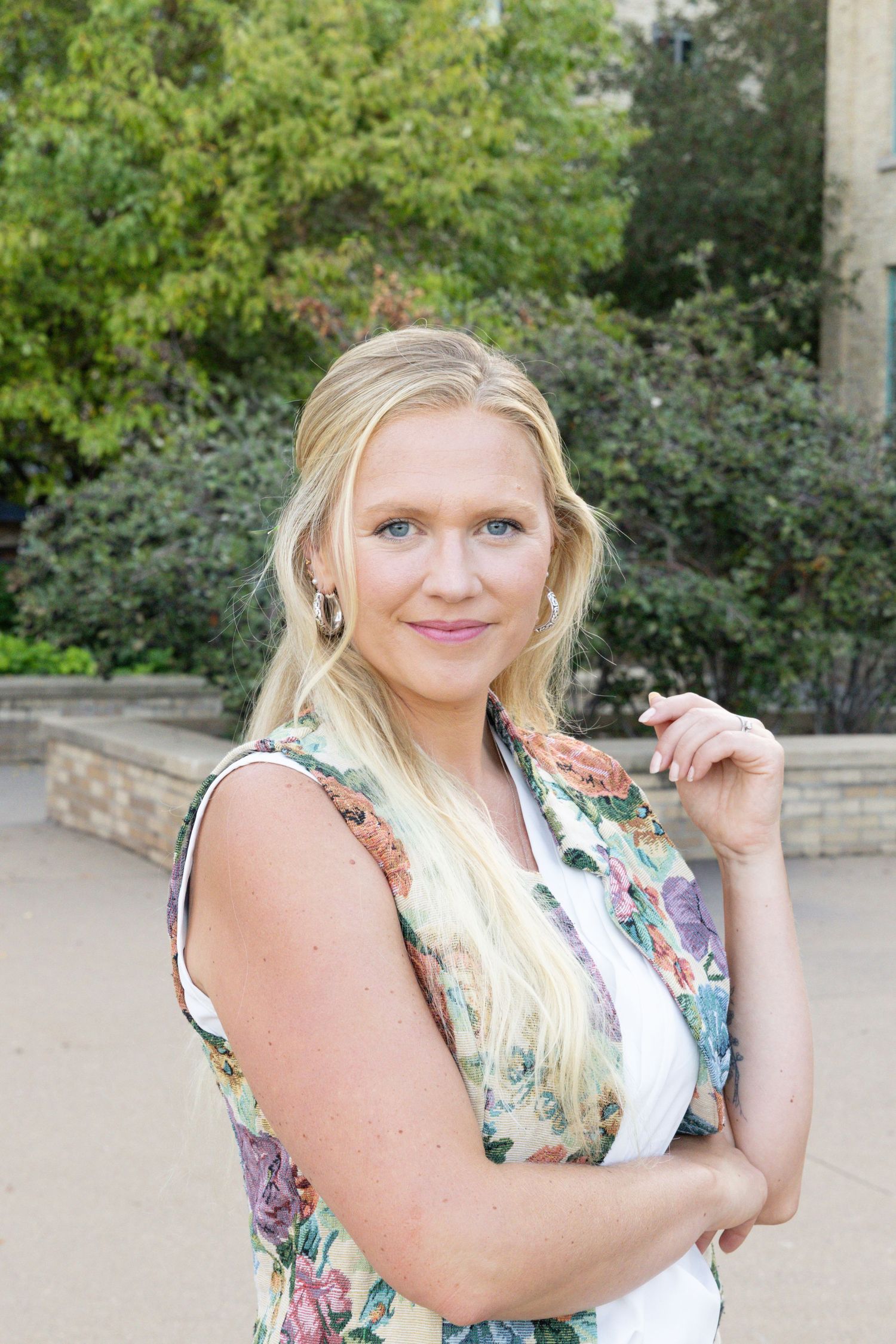 A person in a floral sleeveless top poses outdoors against greenery and stone architecture on a sunny day.