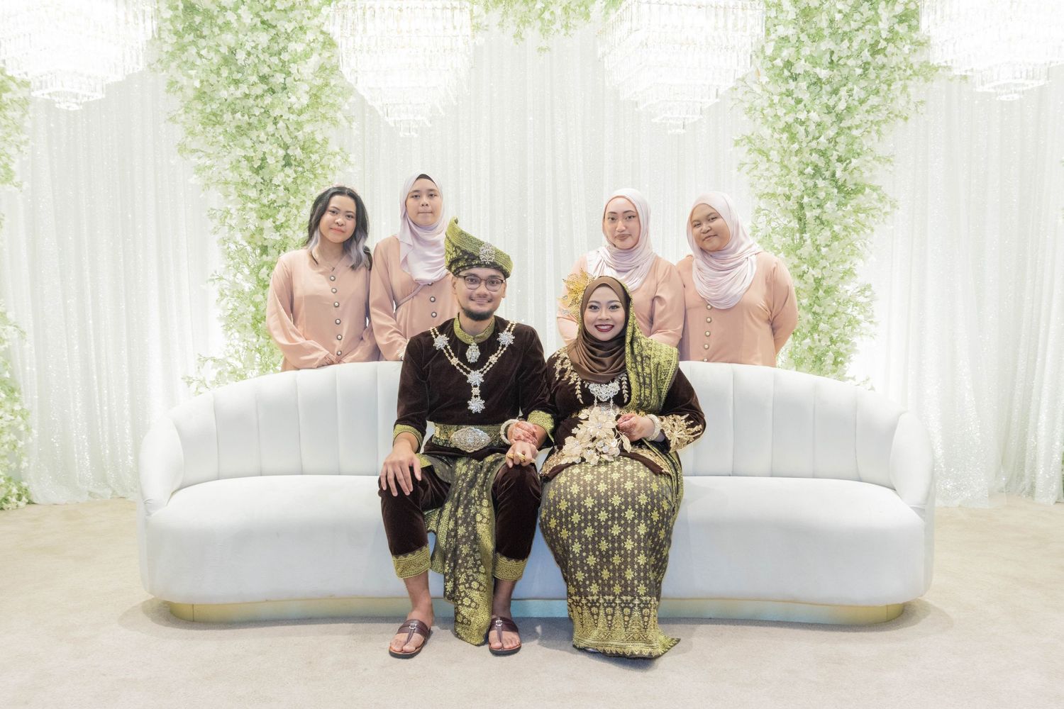 A family group photo on a white couch with traditional Malay formal wear and floral backdrop.