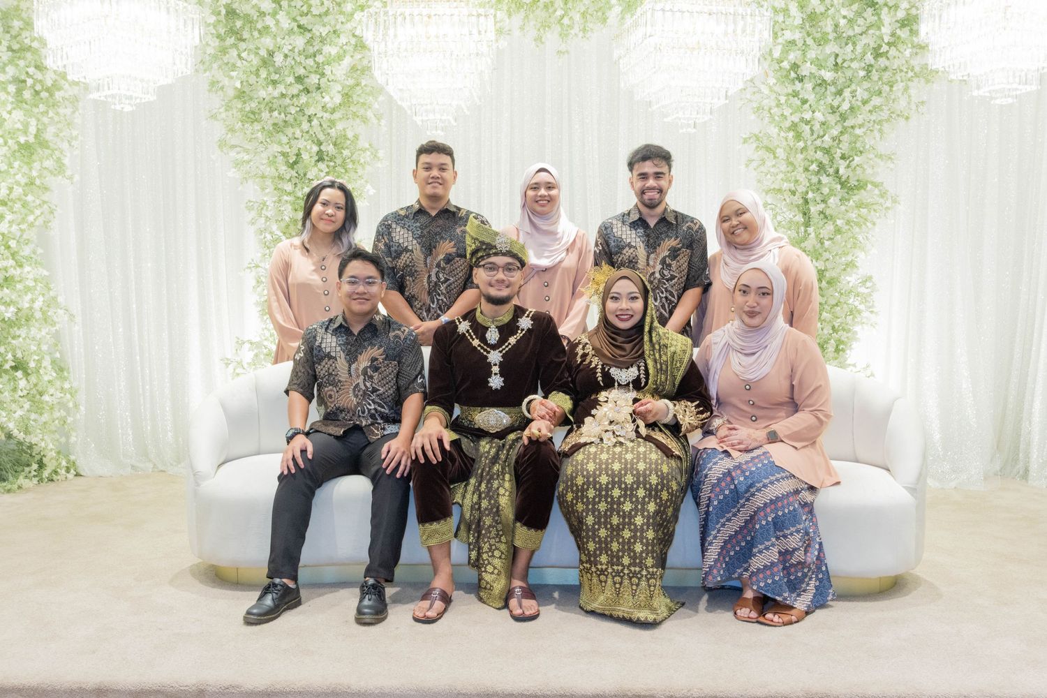 Extended family portrait on white sofa with traditional Malay attire against green floral backdrop.